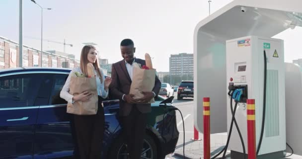 Jóvenes profesionales masculinos que utilizan la computadora en el estacionamiento cerca del supermercado, freelancer. Hombre afroamericano enfocado y joven hermosa mujer, mujer de negocios escribiendo en el ordenador portátil, trabajo remotamente — Vídeos de Stock