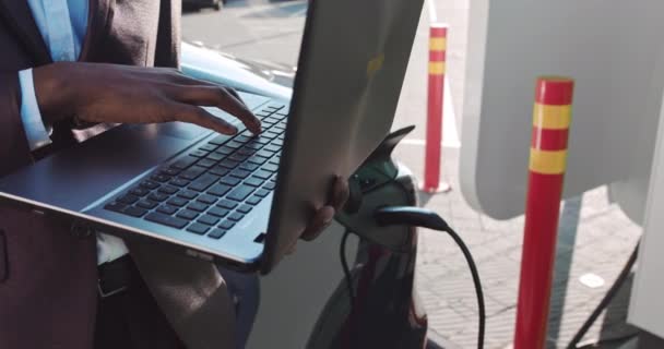 Close-up Macro Shot: Persoon typen op het toetsenbord. Werken, e-mails schrijven. Een Afro-Amerikaanse freelance werknemer houdt een laptop in zijn handen en leunt op een elektrische auto die is opladen. — Stockvideo