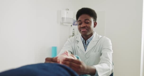 Medico massaggiatore afroamericano. Donna che riceve un massaggio facciale. Il fisioterapista conduce la terapia terapeutica di massaggio ad una giovane donna per ripristinare la forza muscolare nella moderna clinica di riabilitazione — Video Stock
