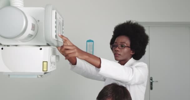 En el hospital, la técnica femenina ajusta la máquina de escáner de rayos X. Hospital moderno con equipos médicos tecnológicamente avanzados y personal profesional. Joven mujer afroamericana Doctor — Vídeos de Stock