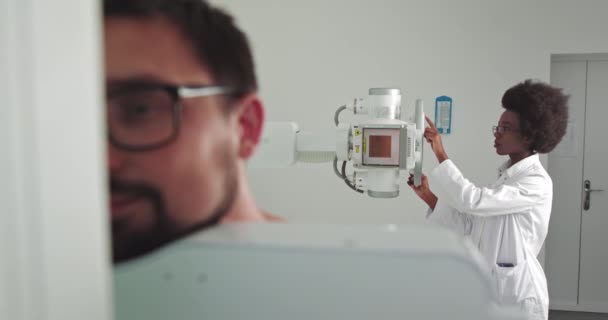 Afrykanka dostosowuje maszynę rentgenowską w gabinecie medycznym. W szpitalu, mężczyzna stojący twarzą do ściany, podczas gdy technik medyczny dostosowuje aparat rentgenowski. Szukanie złamań, klatka piersiowa — Wideo stockowe