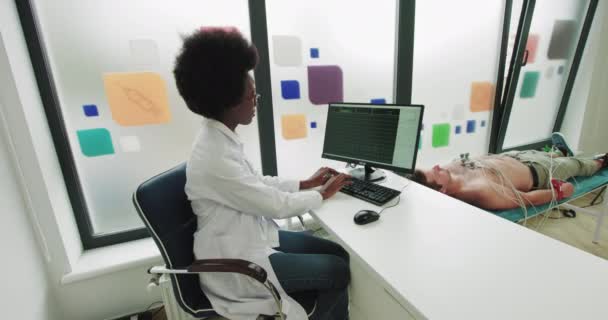 O médico afro-americano está sentado num computador de trabalho que está ligado a um paciente. ECG Patient Health Study. Em laboratório usando eletrodos anexados ao corpo. Monitoramento mostrando ECG e atividade de dados médicos — Vídeo de Stock