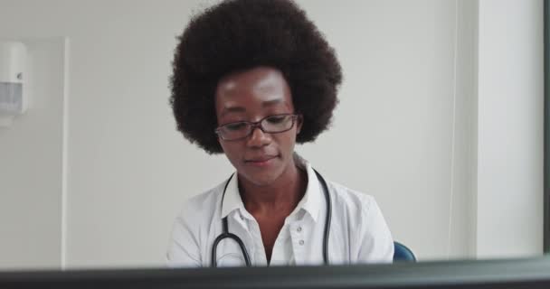 Fermer Portrait of African American Family Medical Doctor in Glasses is in Health Clinic. Médecin noir avec succès en blouse blanche regarde le moniteur et sourit dans le bureau de l'hôpital. — Video