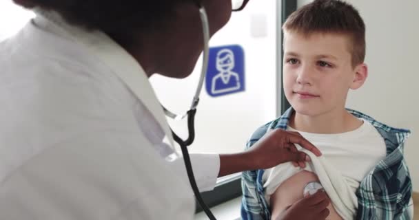 Jovem mulher negra africana em um casaco branco, pediatra exemplifica um menino doente, ouve a respiração usando um fonendoscópio. Doutor e criança. Ambulância. Tiro Médio. Uma criança em uma enfermaria de hospital. — Vídeo de Stock