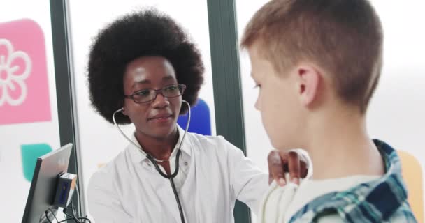 Mooie dokter luistert naar de hartslag van het kind. Jonge vrouw in een witte jas, kinderarts kinderarts onderzoekt een zieke jongen, luistert naar de ademhaling met behulp van een fonendoscoop. Kind in een ziekenhuisafdeling — Stockvideo