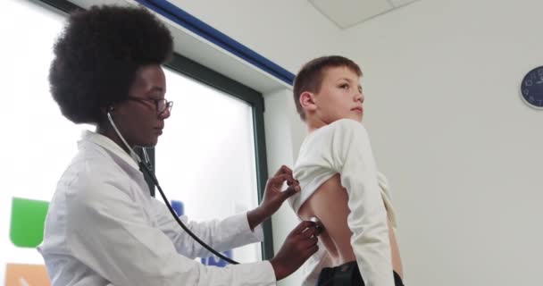 Svart afrikansk kvinna doktor och barn. Vackra doktor lyssnar på barnets hjärtslag. Kvinna i en vit rock, barnläkare undersöker en sjuk pojke, lyssnar på att andas med hjälp av en honendoscope. — Stockvideo