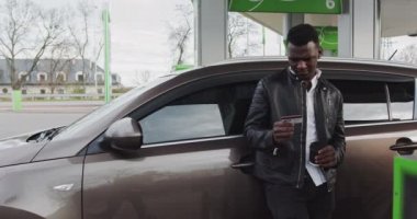 Benzin istasyonunda elinde kahve olan Afrikalı bir adamın portresi. Adamın biri dükkanda indirimli fiyata plastik kart tutuyor ve kamerayı işaret ediyor. Duraklama Benzin İstasyonunda Araba Dolduruluyor