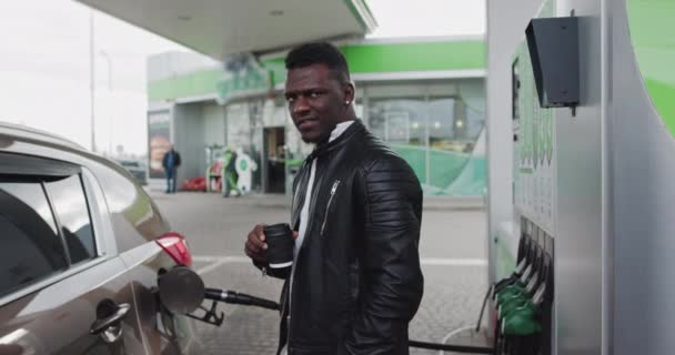 Egy afrikai fickó portréja egy benzinkútnál, kávéval a kezében. Az autó feltöltése utazás közben, megállás a benzinkútnál, férfi kávézás. Üzemanyag, benzinkút, benzinár-koncepció — Stock videók