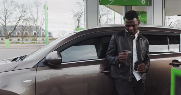 Retrato de um tipo africano num posto de gasolina com café nas mãos. Um homem segura um cartão de plástico com desconto na loja e aponta para a câmera. Reabastecendo o carro enquanto viaja parando a estação GAS — Vídeo de Stock