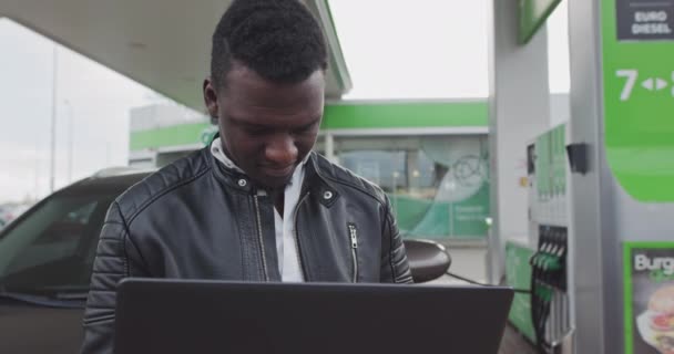 一位非裔美国人自由工作者手里拿着一台笔记本电脑，靠在豪华轿车上。《路上的工作》 ，弗里兰瑟。在加油站的车。燃料、加油站、汽油价格概念. — 图库视频影像