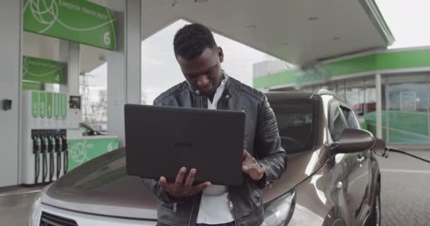 Um trabalhador afro-americano freelancer tem um laptop em suas mãos e se apoia em um carro. Trabalho Freelancer. Carro no posto de gasolina. Combustível, Conceito de Preços da Gasolina. Tradução: ENERGIA DA SUA CIRCULAÇÃO — Vídeo de Stock