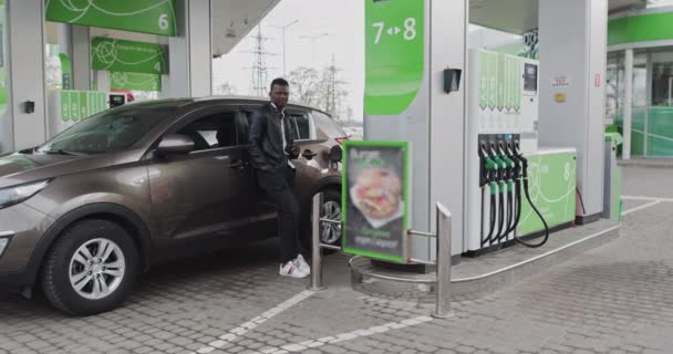 Retrato de um tipo africano num posto de gasolina com café nas mãos. Reabastecendo o carro enquanto viaja, parando na estação GAS, homem bebendo café. Estação de Combustível Tradução: ENERGIA DA SUA CIRCULAÇÃO — Vídeo de Stock