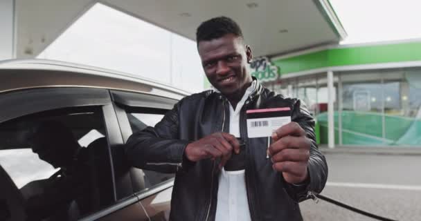 Πορτρέτο ενός Αφρικανού σε βενζινάδικο με καφέ στα χέρια. Ένας άντρας έχει μια πλαστική κάρτα με έκπτωση στο κατάστημα και δείχνει προς την κάμερα. Ανεφοδιασμός του αυτοκινήτου ενώ ταξιδεύετε Σταματώντας το σταθμό του φυσικού αερίου — Αρχείο Βίντεο
