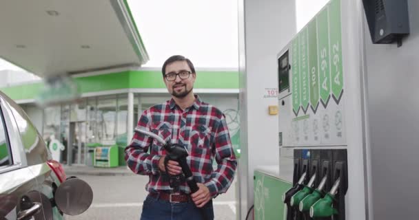 Portret młodego przystojniaka w okularach, stojącego na stacji benzynowej i trzymającego w rękach wtryskiwacz paliwa. Człowiek uśmiechnięty do kamery. Paliwo na stacji benzynowej — Wideo stockowe