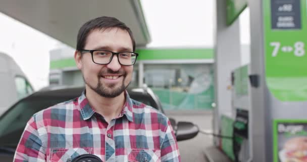 Nahaufnahme eines fröhlichen, gutaussehenden Mannes mit Brille, der neben seinem Auto steht, während er an einer Tankstelle tankt oder Diesel tankt. Porträt des jungen hübschen Kerls. Autotanken. Geschäftsmann blickt auf Kamera — Stockvideo