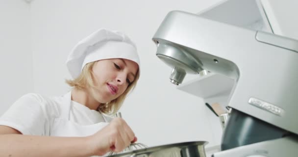 Glückliches hübsches Konditormädchen, das einen elektrischen Schneebesen mit Schlagsahne in der Hand hält, während es einen Kuchen oder Desserts zubereitet, mit moderner Küchenmaschine. Konzept vom Backen, Kochen. Porträt eines Konditors — Stockvideo