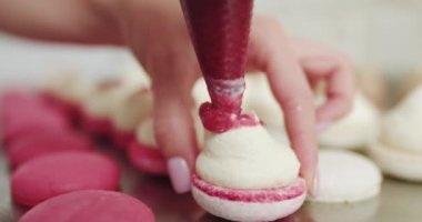 Pembe arka planda pembe reçelle doldurulmuş güzel pembe badem kurabiyeleri. Doğa çörekleri. Kadın pasta şefinin ellerinde beyaz makarna, ganache ve torbadan sıkılmış kırmızı meyve reçeli tutarken.