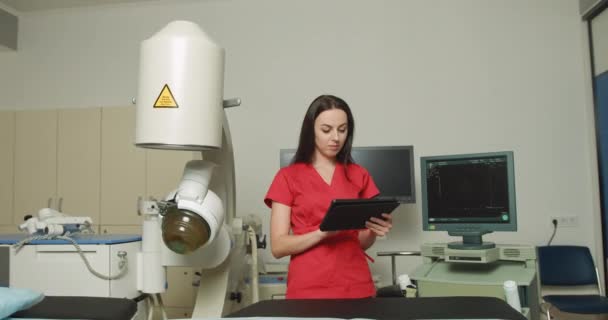 Retrato de cerca de la joven doctora caucásica en uniforme rojo, sosteniendo tableta digital pc, posando cerca de la moderna máquina de litotripter ultrasónico en el centro médico de urología — Vídeo de stock