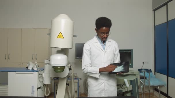 Young African American man doctor in white uniform and eyeglasses, demonstrating digital tablet pc with ultrasound scan, posing near modern ultrasonic lithotripter machine in urology medical center — Stock Video