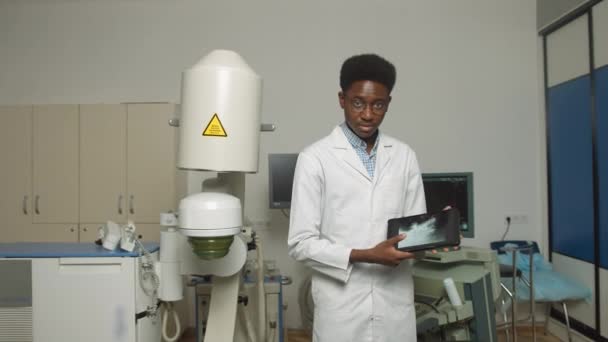 Close up portrait of young African American man doctor in white uniform and eyeglasses, holding digital tablet pc, posing near modern ultrasonic lithotripter machine in urology medical center — Stock Video