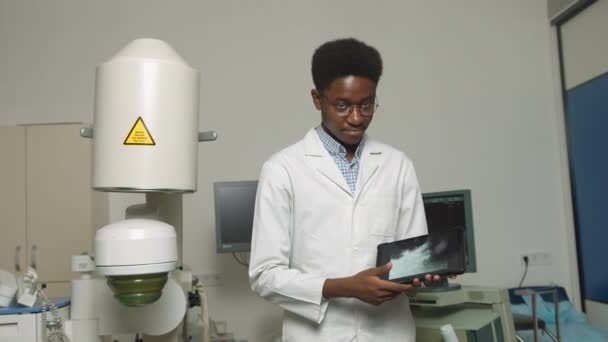 Γιατρός κρατώντας ψηφιακό tablet PC. Κοντινό πορτραίτο του χαμογελαστού όμορφου Αφρικανού γιατρού, που στέκεται μπροστά σε υπερήχους και λιθοτριπτικές μηχανές. Μη επεμβατική θεραπεία λίθων ασφάλειας. — Αρχείο Βίντεο