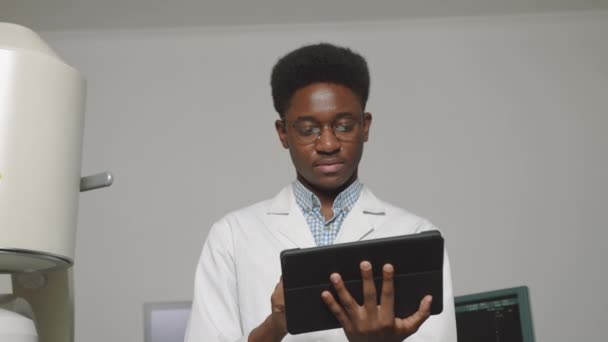 Elinde dijital tablet olan gülümseyen Afrikalı doktorun portresi. Ultrason ve lithotripter makinelerinin önünde duruyor. Güvenlik ihlalsiz taş tedavisi. — Stok video