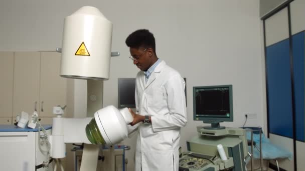 Portait de joven guapo confiado enfocado médico africano, vistiendo uniforme blanco, trabajando en la clínica moderna con máquina de litotripter ultrasónico. Concepto de tratamiento de cálculos renales — Vídeo de stock