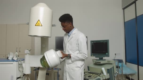 Beyaz üniforma giyen, modern klinikte ultrasonik litotripter makinesiyle çalışan yakışıklı, kendine güvenen genç bir Afrikalı doktor portresi. Böbrek taşı tedavi konsepti — Stok video