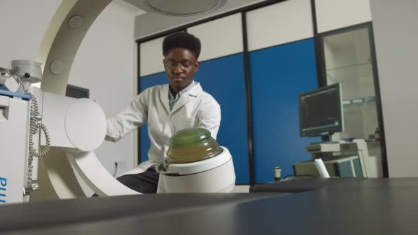 Visão inferior do jovem médico africano confiante em uniforme branco, trabalhando com equipamento litotripter moderno para o tratamento de pedras nos rins. Litotripsia por onda de choque extracorpórea não invasiva — Vídeo de Stock