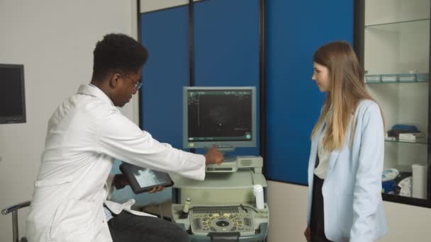 African American Family Medical Doctor în clinica modernă, ținând tableta PC și arătând pacientei sale caucaziene scanarea cu ultrasunete a rinichilor și a organelor interne și explicând modalitățile de tratament — Videoclip de stoc
