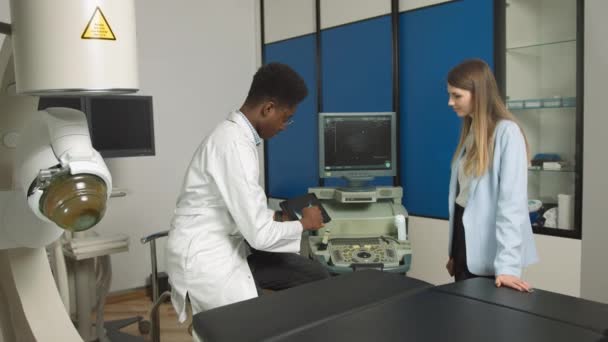 现代诊所的非裔美国家庭医生，手持平板电脑，向她的白人女性病人展示肾和内脏超声扫描，并解释治疗方法 — 图库视频影像