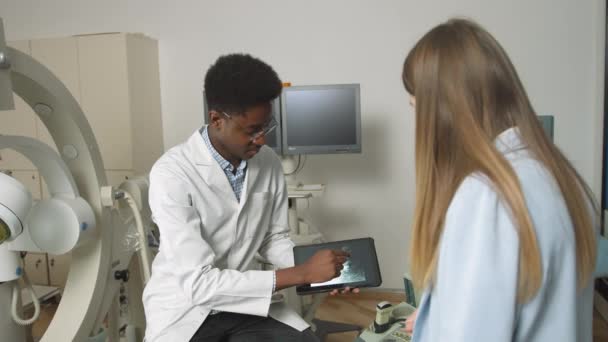 Médico de família afro-americano na clínica moderna, segurando tablet pc e mostrando a sua ultrassonografia paciente caucasiano feminino de rins e órgãos internos e explicando maneiras de tratamento — Vídeo de Stock