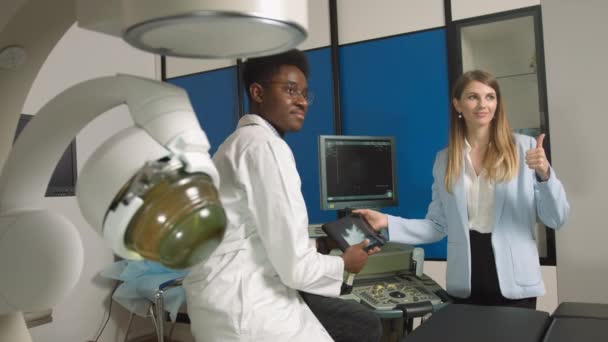 在现代诊所看她的非洲男医生，拿着数字平板电脑进行超声波扫描时，她面带微笑的漂亮女人的画像，摆出相机的姿势。胆道结石治疗和超声检查 — 图库视频影像