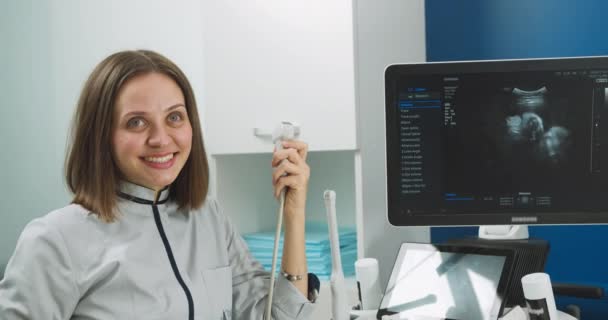 Крупним планом портрет професійної усміхненої молодої жінки-лікаря-сонографа, що сидить біля сучасної машини ультразвукового сканера та тримає перетворювач — стокове відео