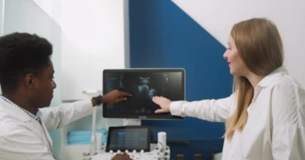Aufnahme einer schönen jungen schwangeren Frau, die auf der Couch im Ultraschall-Untersuchungsraum sitzt und mit ihrem afrikanischen Arzt Rücksprache hält und die Ergebnisse des Ultraschalltests auf dem Bildschirm zeigt — Stockvideo