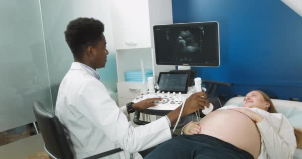 Ultrasound kehamilan. Dokter muda Afrika yang terampil dengan kemampuan tinggi, melakukan pemeriksaan pada perut hamil dengan alat ultrasonografi untuk pasien wanita Kaukasia mudanya — Stok Video