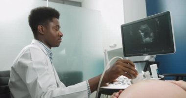 Genç, profesyonel Afrikalı erkek doktor, klinikte hamile, beyaz bir kadının ultrason taramasını yaparken ekrana bakıyor. Doktorların yüzüne odaklan.