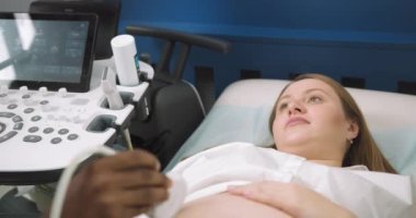 Güzel göbekli hamile sarışın beyaz kadın modern jinekoloji ve doğum kliniğinde ultrason taraması yaptırıyor. Erkek Afrikalı doktorun elleri ultrason taraması yapıyor.