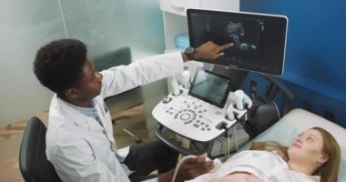 Ultrason konsepti. Beyaz tenli hamile bir kadının kanepede yatarken üst açılı görüntüsü. Bu sırada yüksek yetenekli Afrikalı Amerikalı doktor ultrason taraması yapıyor ve bebeği ekranda gösteriyor.