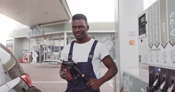 Guapo chico africano joven, trabajador de gasolinera, vistiendo camiseta blanca y mono azul y sosteniendo la pistola de llenado. Vista frontal cerrada. Traducción: ENERGÍA DE SU MOVIMIENTO — Vídeo de stock