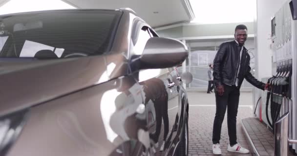 Mladý usmívající se příjemný Afričan, pózující před kamerou, když stojí na benzínce, připraven natankovat své luxusní auto, když si bere plnicí pistoli. Překlad: ENERGIE VAŠE POHYB — Stock video