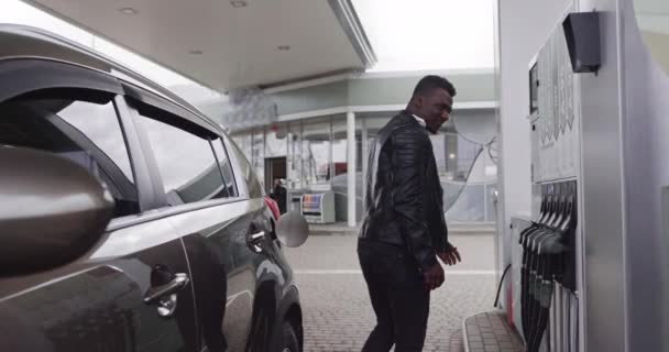 Ung stilig afrikansk skäggig kille i snygg casual outfit tankning sin lyx bil och tittar bort när du står på självbetjäning bensinstation. Översättningar: ENERGI AV DIN MOVEMENT — Stockvideo