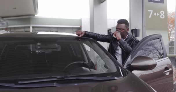 Boldog mosolygós fiatal fekete férfi, tartja el a kávéscsészét, és támaszkodik az autójára, miközben pózol a kamerának a benzinkútnál — Stock videók