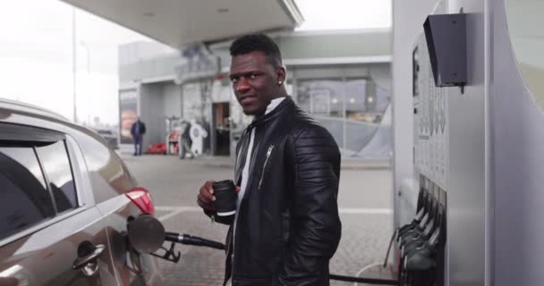 Concepto gasolinera. Relajado soñador joven guapo empresario africano en traje negro, disfrutando de su café para llevar, mientras repostaba su coche de lujo en la gasolinera — Vídeos de Stock