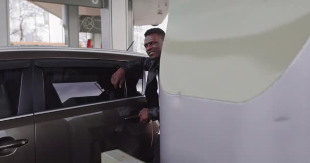 Retrato do jovem e bonito homem afro-americano sorridente, vestindo roupas casuais pretas, olhando para a câmera com café para ir enquanto reabastece o carro no posto de gasolina — Vídeo de Stock