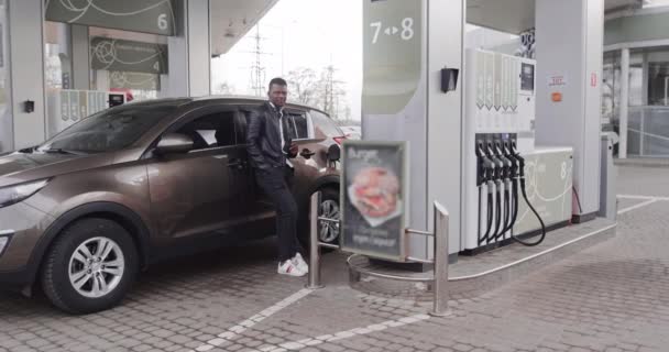 Genç odaklı siyah deri ceketli Afrikalı işadamı, kahvesini içerken modern benzin istasyonunda arabaya yaslanıyor. Çeviri: HAYATININ ENERJİSİ — Stok video