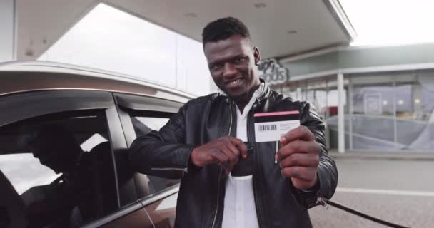 Portret van een vrolijke Afro-Amerikaanse man in casual kleding, die de creditcard aan de camera laat zien, terwijl hij bij het zelfbedieningstankstation staat en zijn moderne auto tankt. Focus op de kaart. Vertaling: ENERGIE — Stockvideo