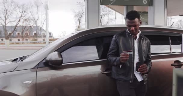 Θολή προσωπογραφία του χαρούμενου Αφροαμερικανού με casual ένδυμα, που δείχνει την πιστωτική του κάρτα στην κάμερα, ενώ στέκεται στο βενζινάδικο self-service και ανεφοδιάζει το σύγχρονο αυτοκίνητό του. Εστίαση στην πιστωτική κάρτα — Αρχείο Βίντεο