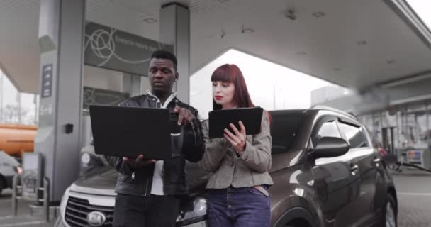 Front view of multiracial male and female friends in casual outfits, working on laptop and tablet, while filling their modern car at gasrol station. Перевод: ENERGY OF YOUR MOVEMENT — стоковое видео