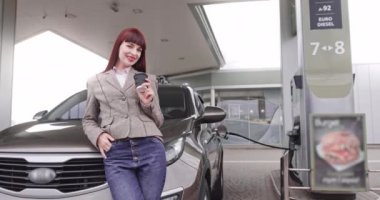 Millet, kahve molası konsepti. Arabasına yaslanıp kahvesini yudumlarken benzin istasyonunda yakıt ikmali yapan, günlük giysiler içinde güzel beyaz bir kız. Çeviri: Enerji Hareketi